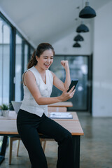 Excited businesswoman raising her fist in celebration, joyfully gazing at her smartphone in a...