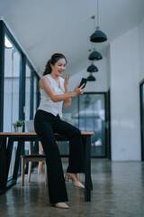 Obraz premium Asian businesswoman sitting on desk waving while making a video call with her smartphone in a modern office, she's wearing elegant clothes and smiling