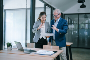 Senior manager and his young female assistant discussing together analyzing financial data document, standing in modern office meeting room with laptop and documents on desk