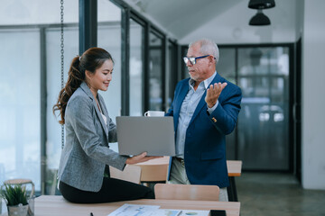 Senior businessman explaining something to young businesswoman holding laptop, sitting on desk with documents, working together in modern office
