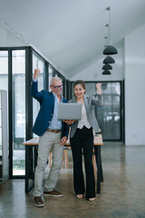 Two happy businesspeople celebrating success while holding a laptop in a modern office, raising their arms in excitement after achieving a great goal