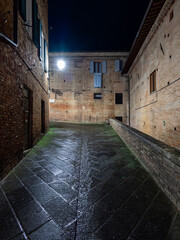A lonely cobblestone street night rain view, Siena. Wet ground reflects light from a lamp and few windows on the brick walls are visible. The street gradually inclines, bordered by a low brick wall.