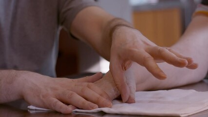 An adult man caregiver assists an elderly woman stroke survivor by gently stimulating and mobilizing her hemiplegic right thumb during a hand therapy session at home kitchen table, on cloth pad.