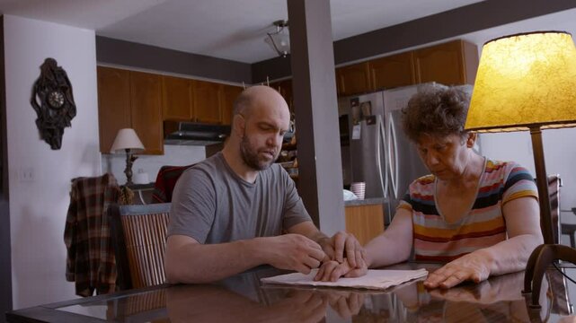 An adult son assists an elderly mother by gently stimulating and mobilizing the fingers of her hemiplegic right hand during a physio therapy session at the kitchen table, on top of a cloth pad. 24fps.