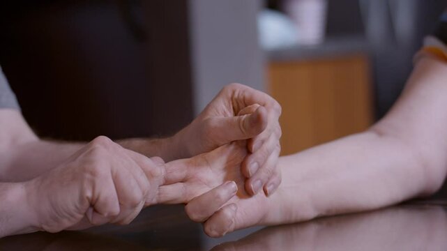Close-up shot of middle-aged son stretching the hemiplegic right hand of elderly mother stroke survivor, on top of a home glass top kitchen table. Focused on thumb, index, and middle fingers. 24fps.