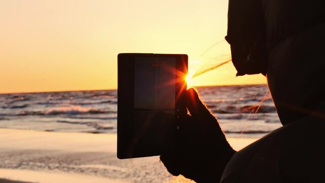 Bright orange sun reflects on tablet screen while person films Baltic Sea sunset