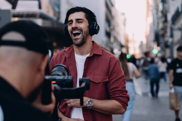 cheerful content creator wearing headphones is being filmed in a busy city street. Male laughs while engaging with the camera, showcasing urban life and digital media culture in a lively atmosphere © BullRun