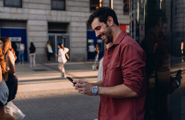 stylish man in a red jacket leans against a glass wall, smiling at his phone. His relaxed posture...