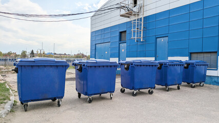 Row of five blue wheeled garbage bins stands outdoors against a blue and white building. Waste management, urban infrastructure