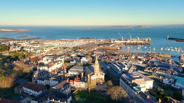 High reveal drone footage of the roof tops of St Peter Port Guernsey in the golden hour featuring Elizabeth College with views across town and the harbour to Herm and Jethou