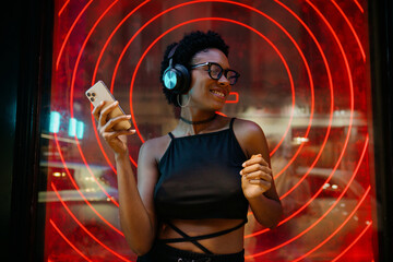 Young stylish Black woman vibing to playlist while streaming music, moving to rhythm with headphones on, neon-lit wall creating bold urban ambiance behind her