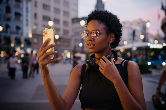 Urban traveler adjusting headphones while chatting on video call, blending fashion and tech as she captures the moment during an evening in a European city