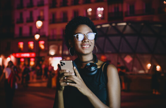 Young woman enjoying nighttime city vibes while holding smartphone, street lights glowing behind, glasses lit by screen, tech-savvy and connected in urban nightlife atmosphere