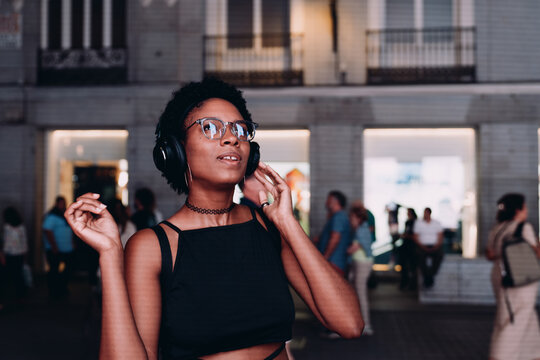Young African American woman enjoying immersive sound through headphones in city at night, pausing to feel rhythm, with look of awe and wonder in her expression