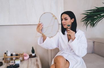 Young woman in white bathrobe using jade roller for morning skincare ritual - natural beauty moment captured with mirror. Peaceful spa-like atmosphere with minimalist cosmetic setup during pastime