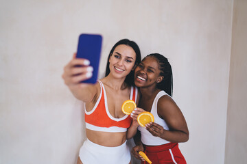Playful cheerful women with fresh orange slices smiling at front mobile camera during together...