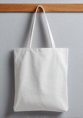 A pristine white cotton tote bag hangs on a light oak wood shelf against a gray wall