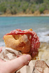 hand hoilding a sandwich with salami in front of the sea