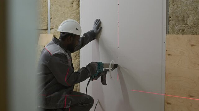 Tracking shot of male African American construction worker in protective gear drilling holes in drywall using power tool guided by laser level during interior wall installation at building site