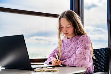 Young calm woman writing ideas in notebook at cafe with laptop, taking notes, journal and pen for planning, sitting at table near window with sky view. Distance study, online education, elearning