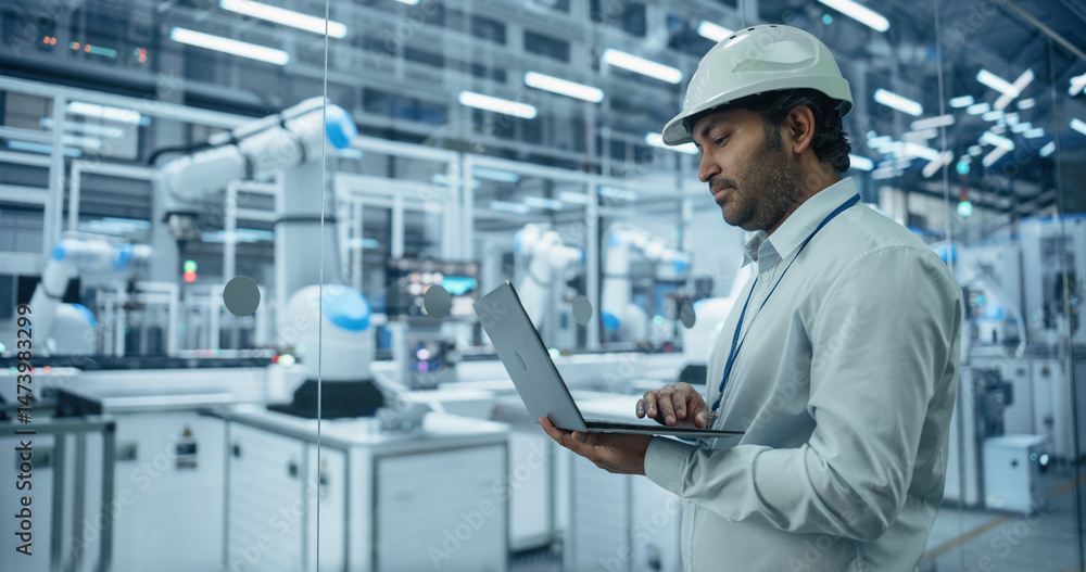 Poster Indian Engineer Working in a High Tech Factory with Robotic Arms Assembling Electronic Components on a Production Line Conveyor Belt. South Asian Man Using Laptop in a Modern Manufacturing Facility
