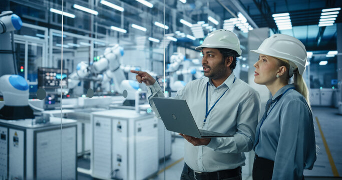 Two Engineers, a South Asian Male and a Caucasian Female, Collaborate Using a Laptop Computer to Optimize Robotic Systems in a Modern Factory, Analyzing Productivity and Automation