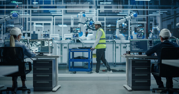 Establishing Shot at an Electronics Factory with Robotic Arms. Diverse Engineers and Developers Working on Desktop Computers. Indian Worker in High Visibility Vest Pushing a Trolley with Equipment