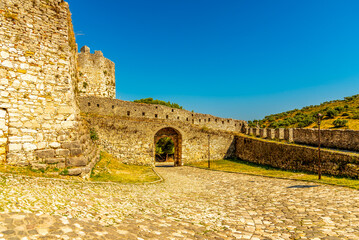 Obraz premium Fortified stone gateway of Berat Castle in Albania with cobblestone courtyard and ramparts atop a sunlit golden hillside under a clear azure sky