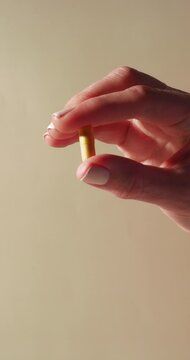 A close-up of a hand gently holding a yellow capsule, possibly a vitamin B complex, cumin, or choline supplement, framed against a neutral beige background with soft daylight highlights.