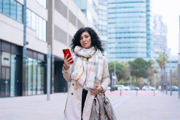 Fototapeta premium Happy caucasian woman standing outdoors in urban setting,wearing coat and scarf,using her red smartphone.She is smiling and relaxed,enjoying a casual moment on a cold day in the city looking at camera