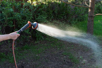 Naklejka premium A person is watering plants with a garden hose in a backyard, creating a spray of water in the sunlight surrounded by greenery and trees.