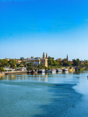 Scenic view of Canal de Alfonso XIII, Seville, Spain, with historic buildings and lush greenery reflecting on calm waters under a clear blue sky.