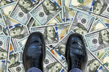 Businessman's black leather shoes standing on scattered hundred dollar bills. Concept of financial...