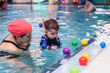 Baby swimming and playing with colorful balls in indoor pool with instructor