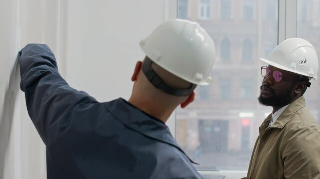 Medium handheld shot of bicultural male interior designers in helmets choosing wall color using swatches in unfinished office space