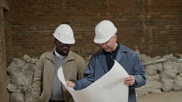 Tracking shot of bicultural male engineers wearing hardhats inspecting blueprint while working on construction site