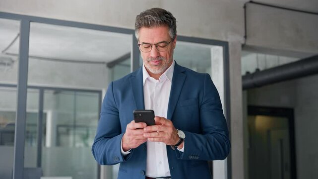 Middle eastern arab or latin senior business manager man ceo using cell phone mobile. Focused Indian mature businessman in suit and eyeglasses holding smartphone standing in office, working on device