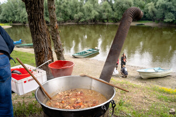People cooking traditional stew by a river with boats in the background during a sunny day