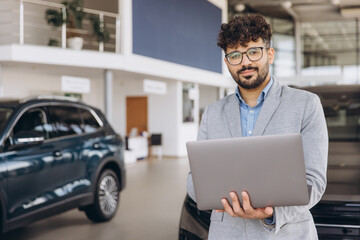 Car salesman holding laptop standing in car dealership showroom