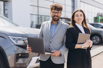 Car salespeople holding laptop and tablet standing in front of new car at dealership