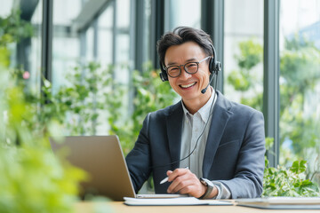 Indian businessman wearing a headset, smiling while talking to a laptop, holding a pen, bright office with large window, fresh greenery outside, modern remote work atmosphere no logo
