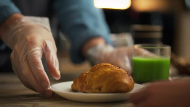 A person wearing gloves serves a perfectly baked croissant on a plate alongside a glass of vibrant green drink. This moment captures the essence of a delightful breakfast experience