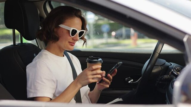 Happy stylish caucasian woman driver in sunglasses serfing social media on smartphone drinking coffee take away, inside car. Girl relaxing chatting with friends. Concept of modern lifestyle