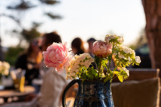 Bouquet de fleurs avec pivoines dans un vase en gros plan sur une table en terrasse au restaurant avec des gens en arri&egrave;re plan au coucher du soleil