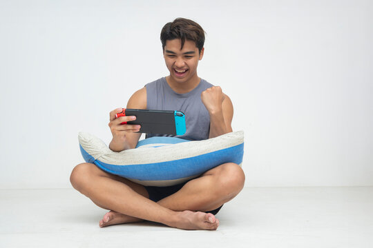 A young Southeast Asian man celebrates with a smile and fist pump while playing a handheld video game console, sitting cross-legged with a pillow. Isolated on a light background.