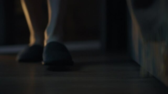 A pair of feet in dark slippers walks across a wooden floor in a softly lit interior. The ambiance suggests a calm and peaceful evening atmosphere, creating a sense of tranquility