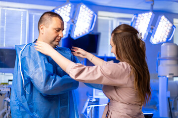 Obraz premium Surgeon prepping for surgery. A female staff member assists a male colleague in putting on surgical attire before a medical procedure in a hospital.