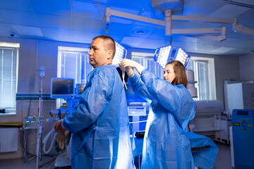 Surgical team readies for surgery. A healthcare worker assists a colleague in wearing surgical...