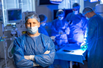 Surgeon in OR during surgery. Surgeon stands confidently with arms crossed while team performs a...