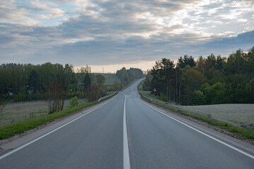 An empty two-lane country road stretches into rolling hills, surrounded by trees and fields under a cloudy morning sky in a peaceful rural landscape